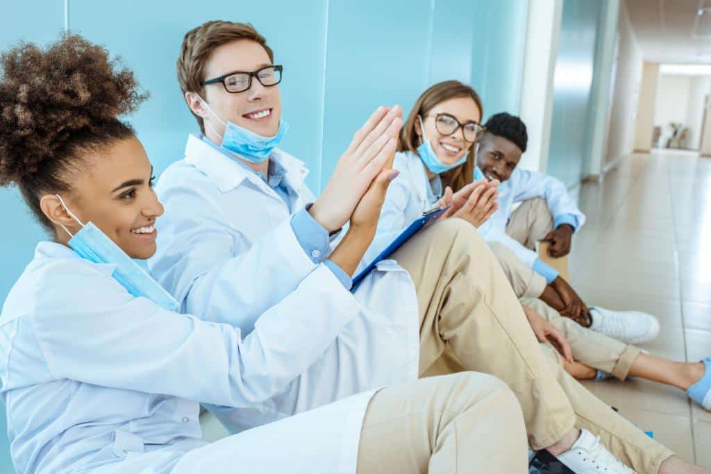 four smiling young medical interns in white robes sitting on a floor in hospital